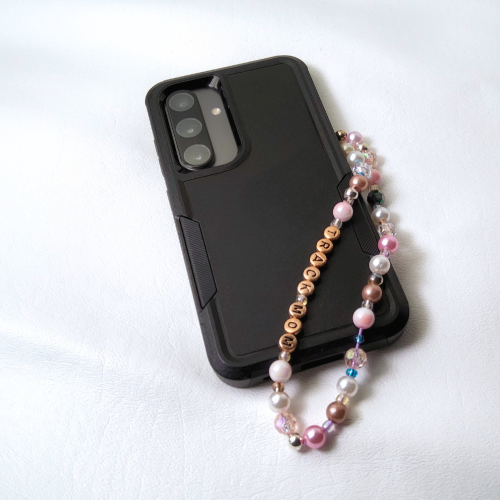 A close-up view of a beaded phone charm. The charm features a mix of white, pink, and rose gold pearl beads, clear crystal beads, and rose gold letter beads spelling 'TRACK MOM'. The perfect gift for every track mom.