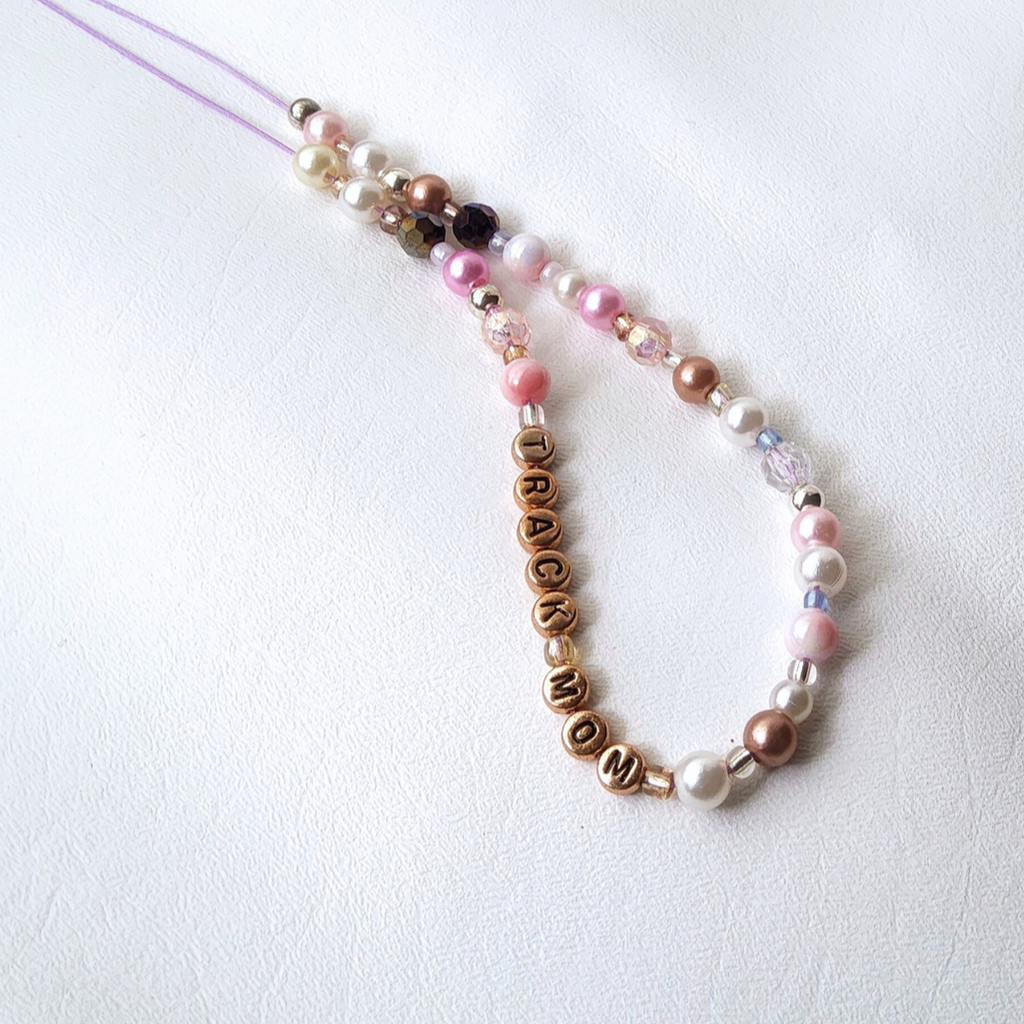 A close-up view of a beaded phone charm. The charm features a mix of white, pink, and rose gold pearl beads, clear crystal beads, and rose gold letter beads spelling 'TRACK MOM'. The perfect gift for every track mom.